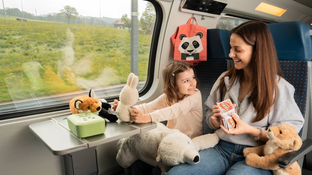 Eine Frau ist mit ihrer Tochter und diversen Kuscheltieren fröhlich im Zug unterwegs. 