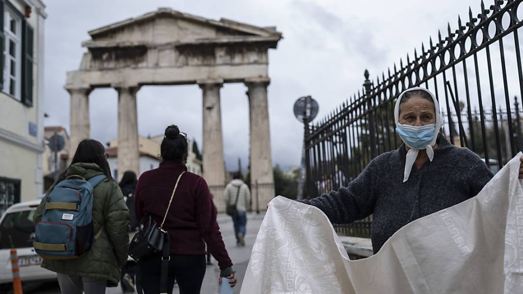 Eine Frau mit Mund-Nasen-Schutz verkauft eine Decke vor dem Tor der antiken römischen Agora in Plaka, einem Stadtteil von Athen. Die griechische Gesundheitsbehörde hat von Montag auf Dienstag 21 657 Corona-Neuinfektionen registriert. Foto: Petros Giannakouris/AP/dpa