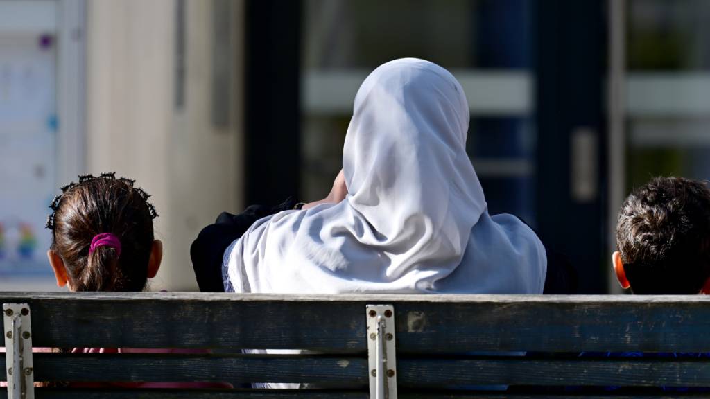 Eine Frau mit Kopftuch sitzt vor einer Schule am ersten Schultag des neuen Schuljahres auf einer Bank zwischen zwei Kindern. Foto: Roland Schlager/APA/dpa