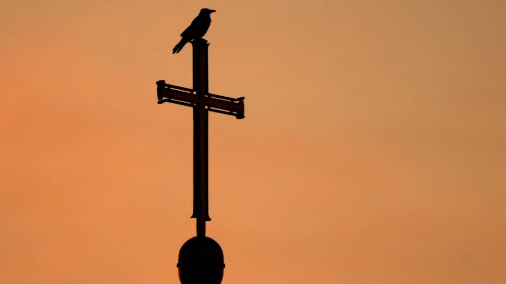 ARCHIV - Ein Kreuz auf einem Kirchturm in der Region Hannover zeichnet sich vor dem vom Sonnenaufgang rötlich verfärbten Morgenhimmel ab. Foto: Julian Stratenschulte/dpa