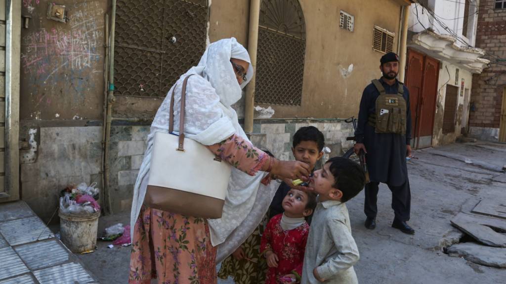 Ein Polizist steht Wache, während eine Gesundheitshelferin (l) einem Kind in einem Wohnviertel in Peshawar, Pakistan, einen Polio-Impfstoff verabreicht. Foto: Muhammad Sajjad/AP/dpa