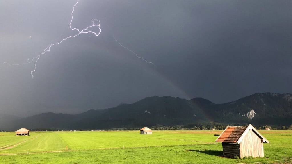 Ein Blitz zuckt über den bayerischen Voralpen, während über einer Weide ein Regenbogen erkennbar ist. +++ dpa-Bildfunk +++
