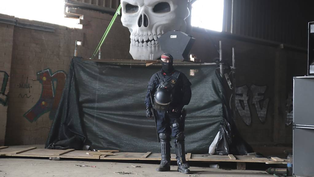 Ein Polizeibeamter der Mossos d'Esquadra steht in einer Lagerhalle vor einem riesigen Totenkopf, während vor Ort ein Rave aufgelöst wird.