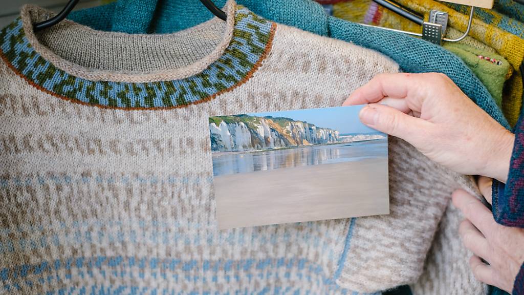 Die Textildesignerin Anne-Susanne Gueler hält in ihrem Werkstattladen «hand-werk» ein Foto aus der Normandie neben einen von ihr gestrickten Pullover. Gueler lässt sich häufig von Landschaften inspirieren. Foto: Ole Spata/dpa