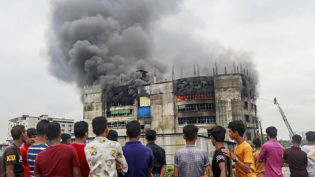 Menschen sehen zu, wie während des Brands in einer Fabrik Rauch von dem Gebäude aufsteigt. Bei einem Brand in einer Lebensmittel- und Getränkefabrik außerhalb der bangladeschischen Hauptstadt Dhaka sind mindestens 52 Menschen getötet worden. Foto: Suvra Kanti Das/ZUMA Wire/dpa
