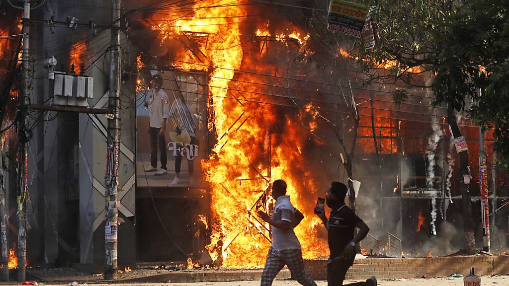 dpatopbilder - Männer rennen an einem Einkaufszentrum vorbei, das von Demonstranten während einer Kundgebung gegen Premierministerin Hasina und ihre Regierung angezündet wurde. Foto: Rajib Dhar/AP/dpa