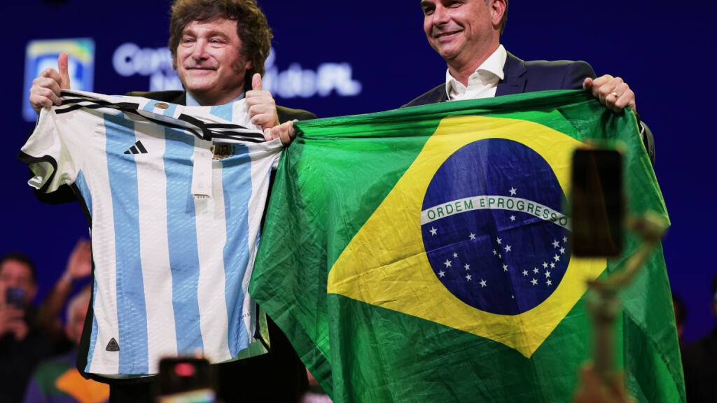 Der brasilianische Senator Flavio Bolsonaro (r) hält eine Nationalflagge, während der argentinische Präsident Javier Milei ein argentinisches Trikot in der Hand hält - dies geschah auf dem Parteitag der Liberalen Partei, bei dem Bolsonaro als Präsidentschaftskandidat der Partei bestätigt wurde. Foto: Ettore Chiereguini/AP/dpa