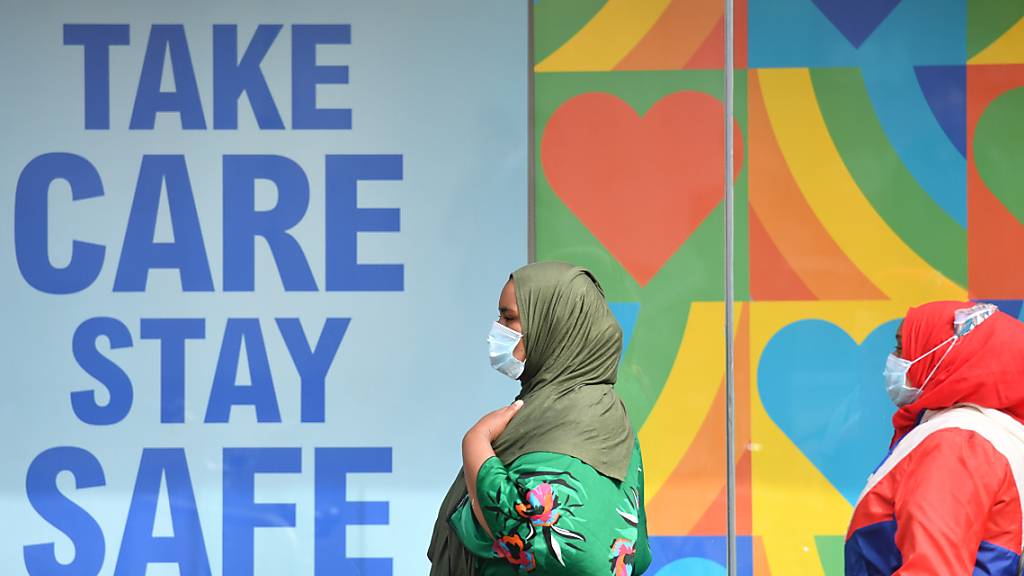 Frauen mit Kopftuch und Mundschutz gehen in einem Stadtzentrum in Großbritannien an einem Plakat vorbei, das an die Corona-Schutzregeln erinnert: «Take care, stay safe». Foto: Jacob King/PA Wire/dpa