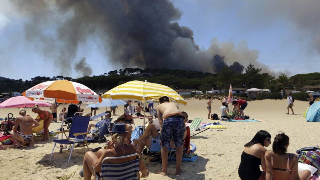 Menschen am Strand von Le Lavandou an der französischen Riviera - im Hintergrund steigen schwarze Rauchwolken in den Himmel. Wegen den Waldbränden in Südfrankreich mussten bereits 10'000 Menschen evakuiert werden.