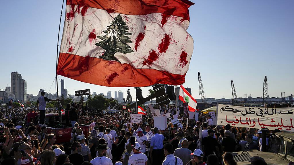 Menschen schwenken eine riesige libanesische Nationalfahne, während sie sich vor dem Hafen versammeln. Am dritten Jahrestag der verheerenden Explosion im Hafen der libanesischen Hauptstadt Beirut haben hunderte Menschen am Unglücksort demonstriert. Foto: Hassan Ammar/AP/dpa