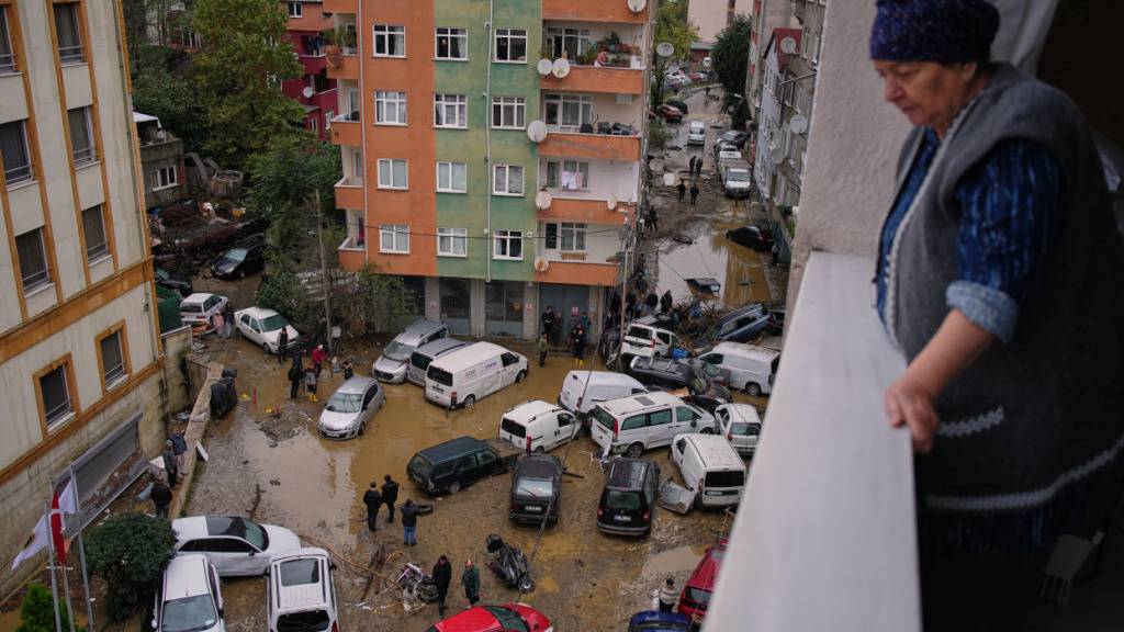 Eine Frau schaut von ihrem Balkon auf Anwohner, die zwischen umgestürzten Fahrzeugen und verstreuten Trümmern umher gehen, nachdem heftige Regenfälle zu Überschwemmungen geführt haben. Foto: Emrah Gurel/AP/dpa