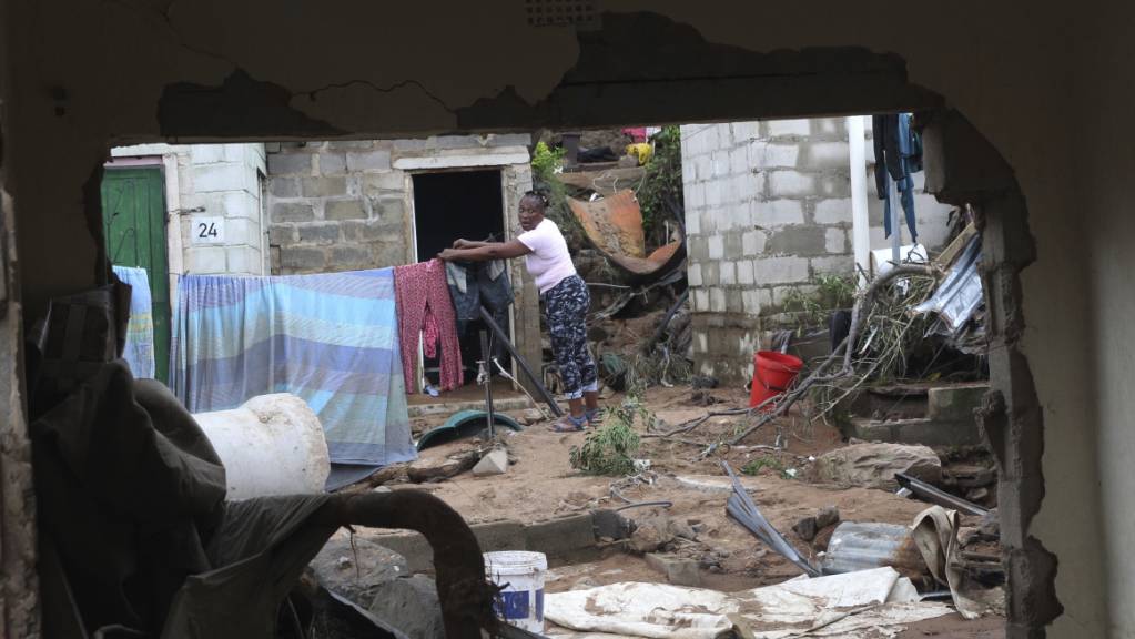 Eine Frau schaut durch eine beschädigte Wand hindurch, während sie ihre Wäsche an einer behelfsmäßigen Wäscheleine aufhängt. In Südafrika kämpfen die Menschen mit den dramatischen Folgen einer Unwetter-Katastrophe mit knapp 400 Toten. Foto: Str/AP/dpa