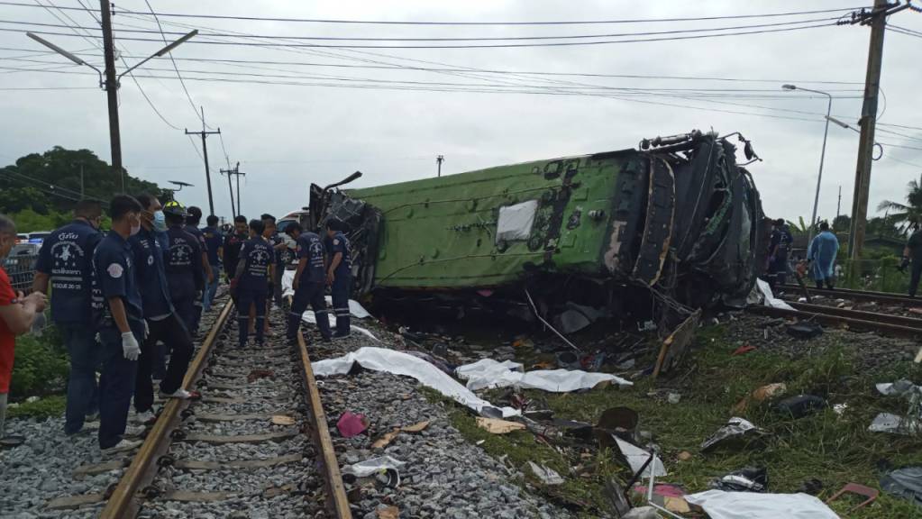 Rettungskräfte stehen an einem beschädigten Zug und neben mit weissen Tüchern bedeckten Leichen. Mindestens 17 Menschen sind am Sonntag in Thailand beim Zusammenstoß eines Busses mit einem Zug ums Leben gekommen. Weitere 30 Menschen wurden bei dem Unfall verletzt. Foto: Uncredited/Daily News/AP/dpa