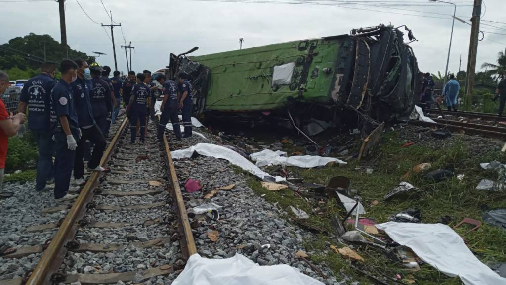 Rettungskräfte stehen an einem beschädigten Zug und neben mit weißen Tüchern bedeckten Leichen. Mindestens 17 Menschen sind am Sonntag in Thailand beim Zusammenstoß eines Busses mit einem Zug ums Leben gekommen. Weitere 30 Menschen wurden bei dem Unfall verletzt. Foto: Uncredited/Daily News/AP/dpa