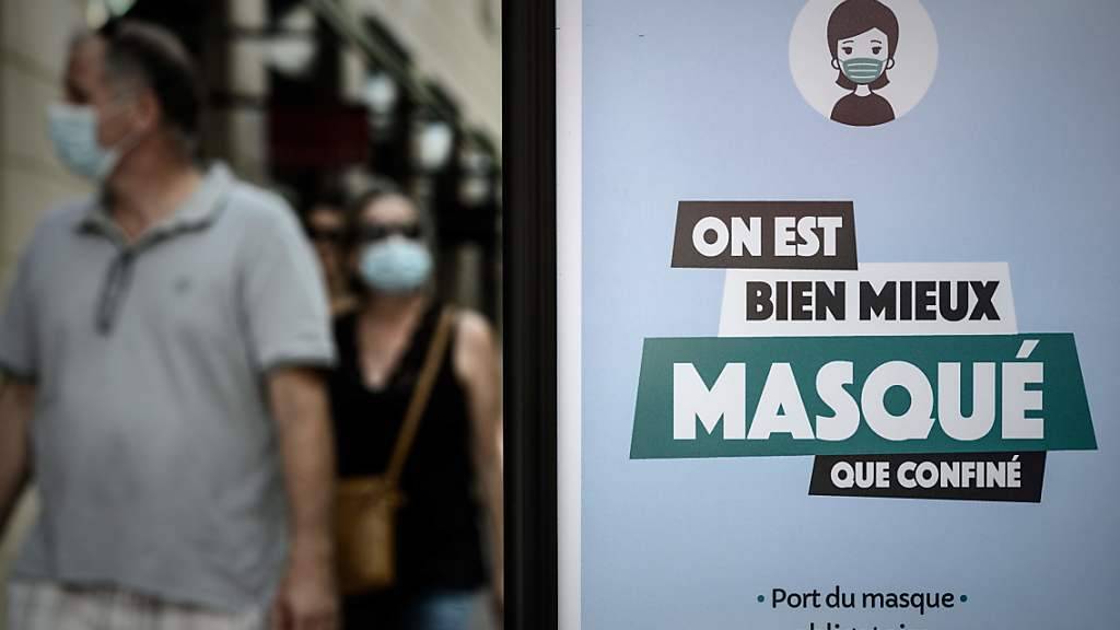 Passanten sind hinter einem Plakat mit der Aufschrift «On est bien mieux masqué que confiné» (Lieber maskiert als eingesperrt) zu sehen. Foto: Philippe Lopez/AFP/dpa