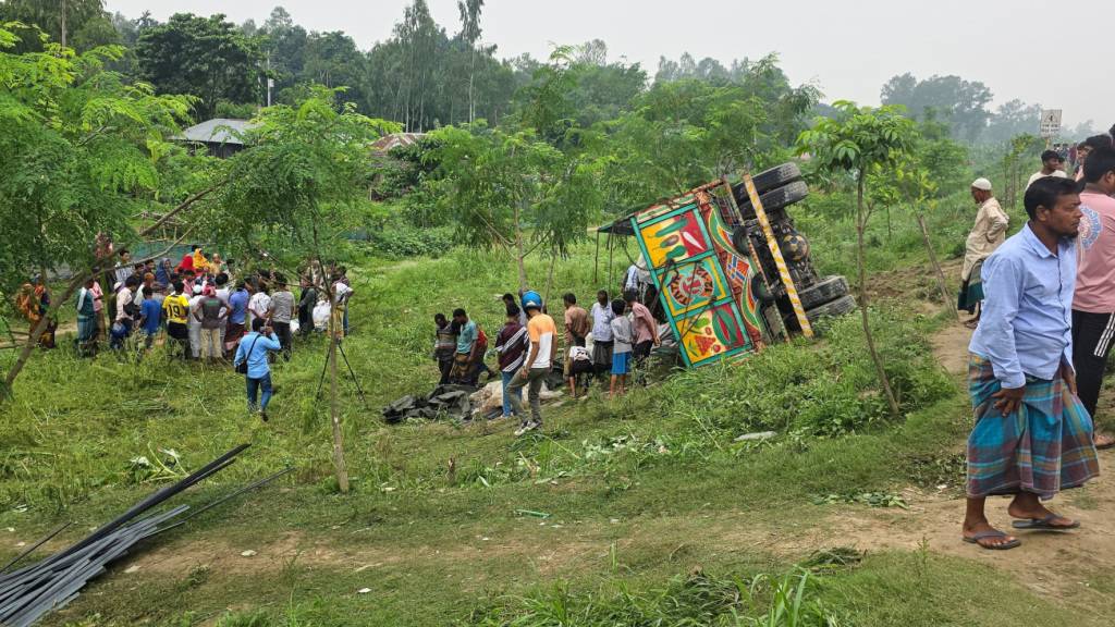 Menschen schauen zu, nachdem ein mit Eisenstangen und zusätzlichen Passagieren beladener Lastwagen auf einer Straße umgestürzt ist. Foto: Sohel Talukder/AP/dpa