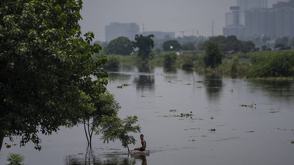 Ein Junge sitzt auf einem Schild in der Nähe einer Wohnkolonie, die vom Hochwasser des Flusses Hindon überschwemmt wurde. Foto: Altaf Qadri/AP/dpa