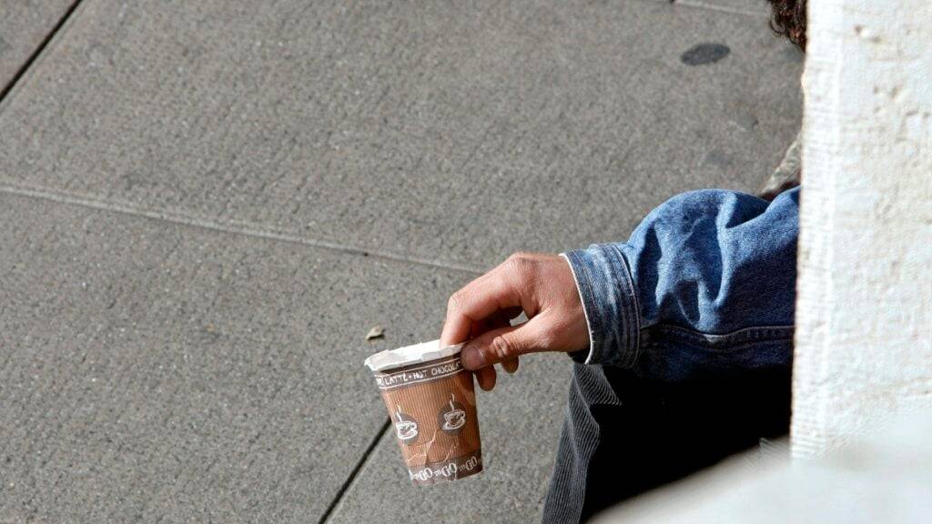 Ein Mann (von hinten) hält einen kaputten Kaffeebecher. Er sitzt auf dem Betonboden.