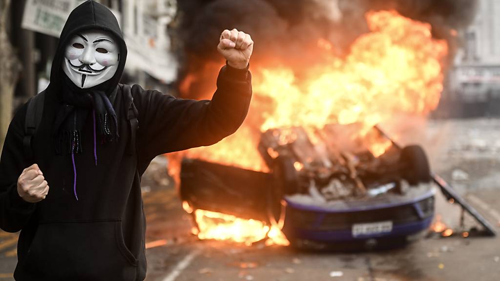 Ein Demonstrant, der eine Anonymus-Maske trägt, steht vor einem brennenden Wagen während eines Protests gegen ein Reformvorhaben der ultraliberalen Regierung von Präsident Milei, das im Kongress debattiert wird. Foto: Fernando Gens/dpa