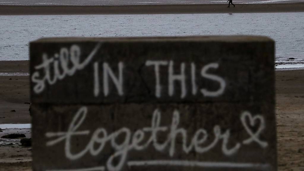 Ein Schild steht am Burrow Beach von Sutton bei Dublin, an dem eine Person allein bei Ebbe spazieren geht. Auf dem Schild ist zu lesen «Wir halten immer noch zusammen». Foto: Brian Lawless/PA Wire/dpa