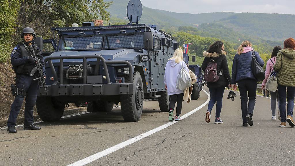 Ein kosovarischer Soldat hält eine Waffe in der Hand und steht auf einer Straße in der Nähe des Grenzortes Jarinje, während Personen an ihm vorbeigehen. Zwischen dem Kosovo und Serbien ist es wegen einer neuen Verordnung über die Zulässigkeit von Kfz-Kennzeichen zu Spannungen gekommen. Foto: Visar Kryeziu/AP/dpa