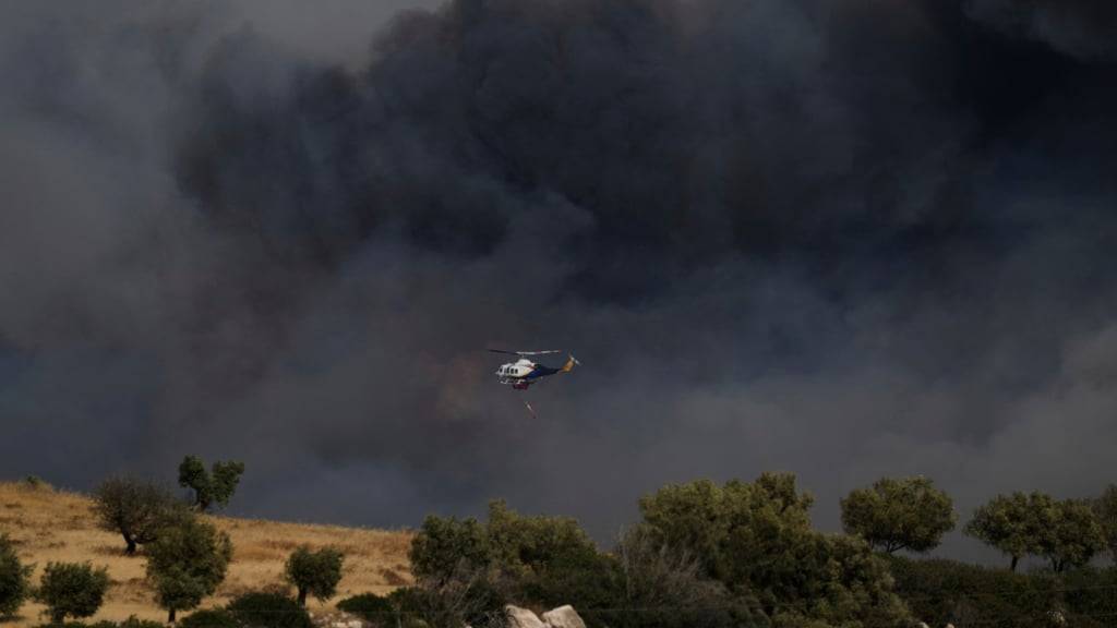 Ein Hubschrauber fliegt durch den Rauch eines Brandes in Keratea, einem Außenbezirk von Athen. Foto: Thanasis Stavrakis/AP/dpa