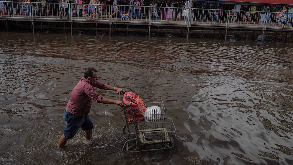 Ein Mann schiebt einen Einkaufswagen durch eine überschwemmte Straße, während Passanten auf provisorisch über dem Wasser angelegten Gehwege spazieren. Foto: Lucas Silva/dpa