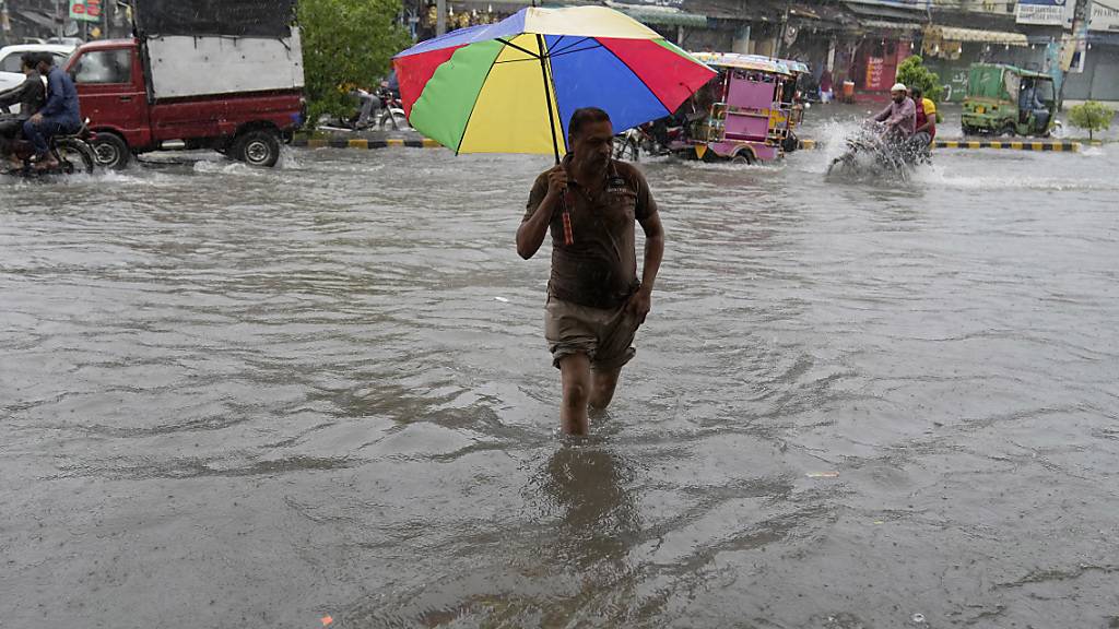 Ein Mann überquert eine überschwemmte Straße, während er sich mit einem Regenschirm schützt. Schwere Monsunregenfälle haben in Pakistan nach offiziellen Angaben bereits mehrere Todesopfer gefordert. Foto: K.M. Chaudary/AP/dpa