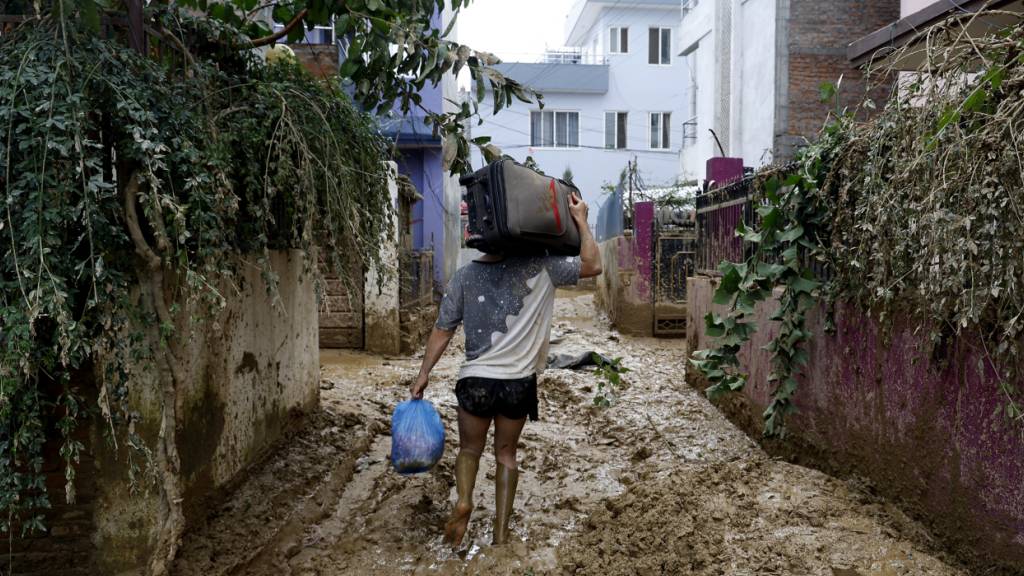 Ein Mann läuft durch eine schlammige Gasse und trägt Habseligkeiten aus seinem Haus nach einer Überschwemmung, die durch starke Regenfälle verursacht wurde. Foto: Gopen Rai/AP