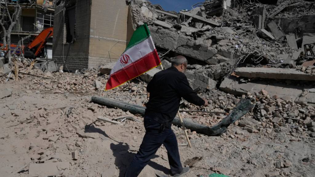 Ein Mann trägt eine iranische Flagge und legt sie auf die Trümmer einer Polizeieinrichtung, die während der amerikanisch-israelischen Militäraktion  getroffen wurde. Foto: Vahid Salemi/AP/dpa