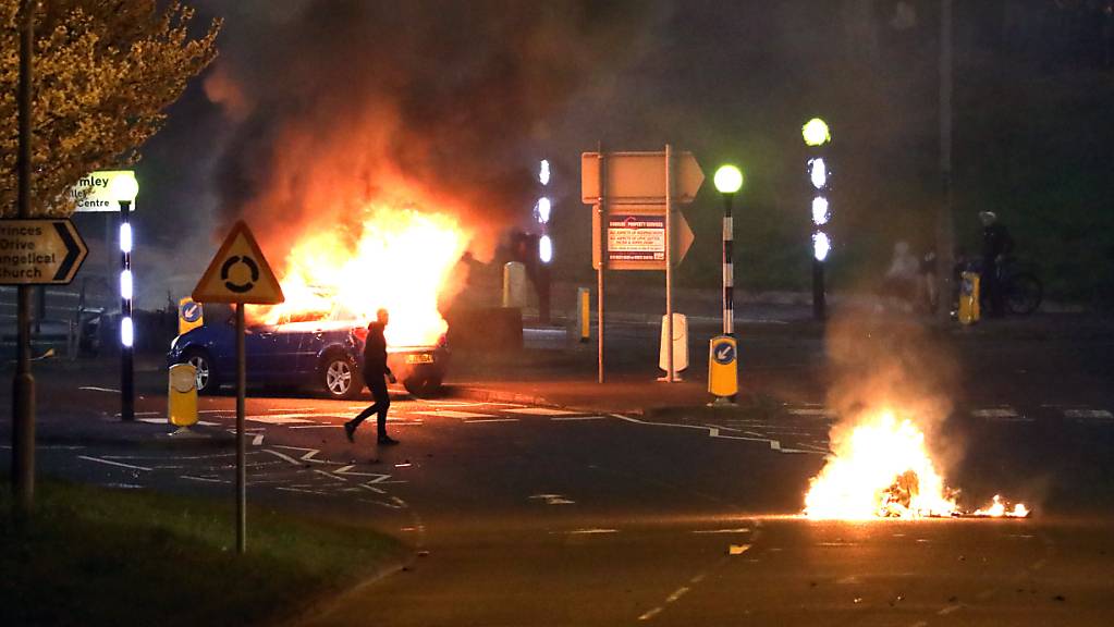 Ein Mann geht an einem brennenden Auto vorbei. Bei Protesten im nordirischen Belfast sind mehrere Polizisten verletzt worden. Foto: Peter Morrison/PA Wire/dpa