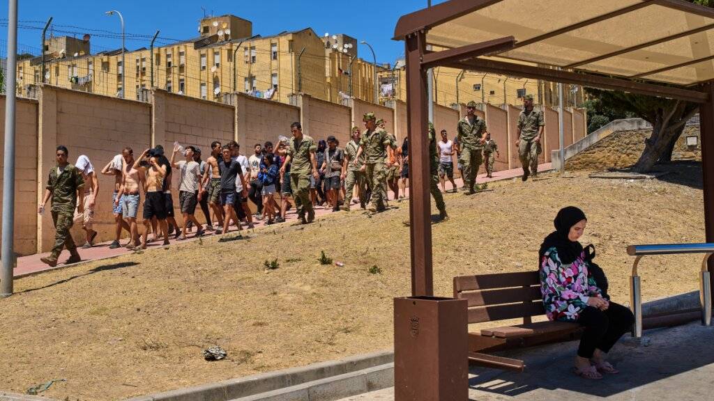 dpatopbilder - Eine Einwohnerin von Ceuta sitzt an einer Bushaltestelle, während Migranten, die nach Spanien eingereist sind, von spanischen Soldaten in Richtung der spanisch-marokkanischen Grenze eskortiert werden, um von der spanischen Enklave Ceuta nach Marokko zurückgebracht zu werden. Foto: Bernat Armangue/AP/dpa