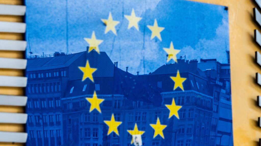 ARCHIV - Drei Fahnen der Europäischen Union und gegenüberliegende Gebäude spiegeln sich im EU-Logo an der Außenfassade des Berlaymont-Gebäudes. (zu dpa: «EU-Kommission will von USA Klarheit zu Zöllen») Foto: Alicia Windzio/dpa