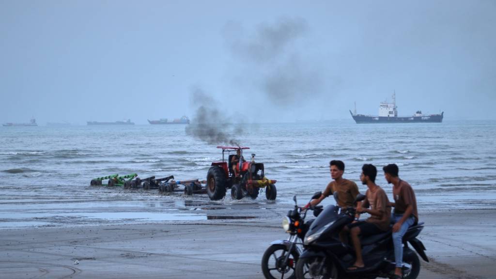 ARCHIV - Junge Männer fahren mit Motorrädern am Ufer entlang, während ein Traktor Geräte aus dem Wasser zieht und Handelsschiffe in der Straße von Hormuz vor Anker liegen. Foto: Razieh Poudat/ISNA/AP/dpa