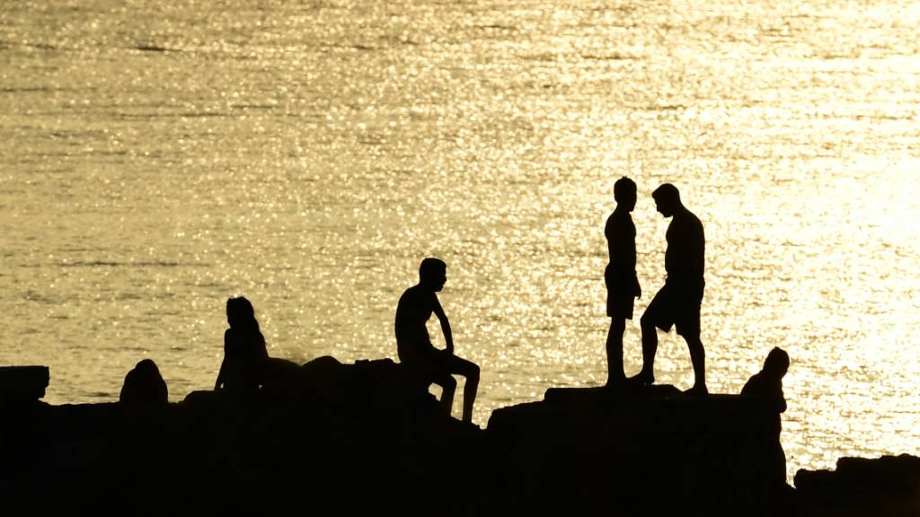 Menschen stehen auf Felsen in der Nähe eines Strandes im Vorort Kavouri. Die extreme Hitzewelle in Griechenland lässt nicht locker. Seit über einer Woche klettern die Temperaturen für mehrere Stunden täglich auf über 40 Grad. Foto: Michael Varaklas/AP/dpa