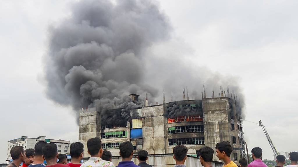 Menschen sehen zu, wie während des Brands in einer Fabrik Rauch von dem Gebäude aufsteigt. Bei einem Brand in einer Lebensmittel- und Getränkefabrik außerhalb der bangladeschischen Hauptstadt Dhaka sind mindestens 52 Menschen getötet worden. Foto: Suvra Kanti Das/ZUMA Wire/dpa