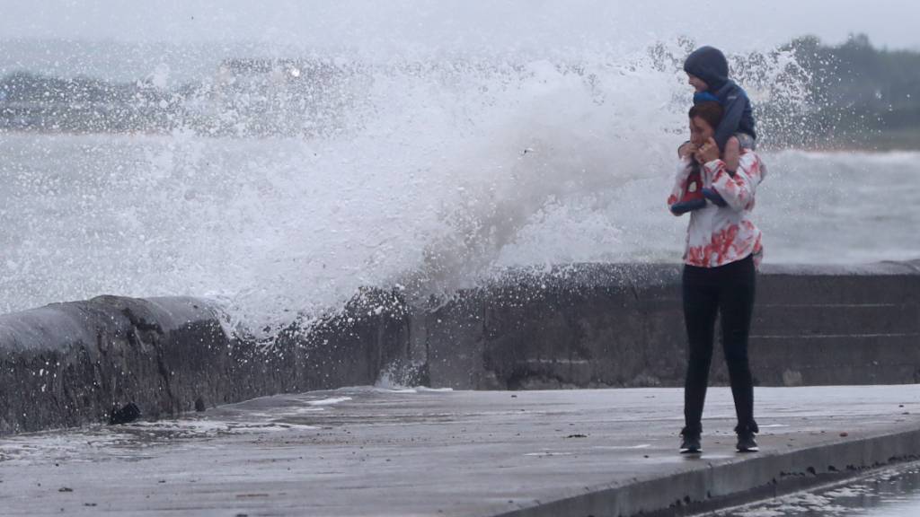 Eine Welle tritt über das Ufer während eine Frau mit einem Kind auf den Schultern an der Strandpromenade geht. Sturmtief «Ellen» trifft auf die Küste Irlands. Foto: Niall Carson/PA Wire/dpa