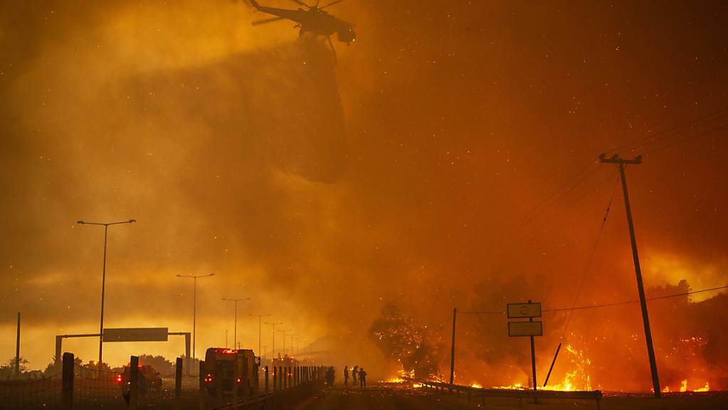In der griechischen Kleinstadt Afidnes stehen Feuerwehrleute auf einer Straße und reinigen einen Feuerwehrschlauch, während im Hintergrund ein Hubschrauber Wasser über einem Waldbrand abwirft. Die dramatischen Waldbrände in Griechenland und der Türkei dauern unvermindert an. Foto: Angelos Tzortzinis/dpa