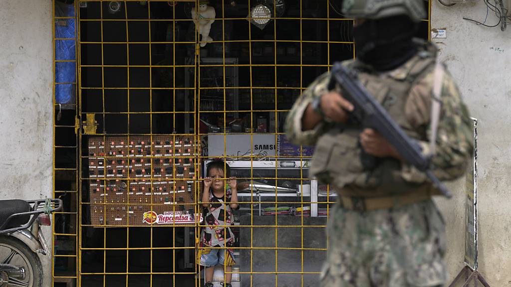 Ein Junge schaut hinter den Gitterstäben eines geöffneten Elektronikgeschäfts hervor, während ein Soldat an einem von der Armee eingerichteten Sicherheitskontrollpunkt Wache steht. Kurz vor der Präsidentenwahl in Ecuador ist ein weiterer Lokalpolitiker im Nordwesten des Landes getötet worden. Foto: Martin Mejia/AP/dpa