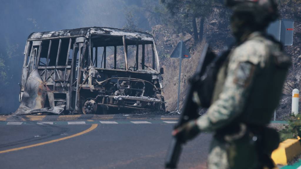 ARCHIV - Ein Soldat steht neben einem ausgebrannten Bus, der in Mexiko nach dem Tod eines Kartell-Anführers in Brand gesetzt wurde. Foto: Armando Solis/AP/dpa/Symbolbild