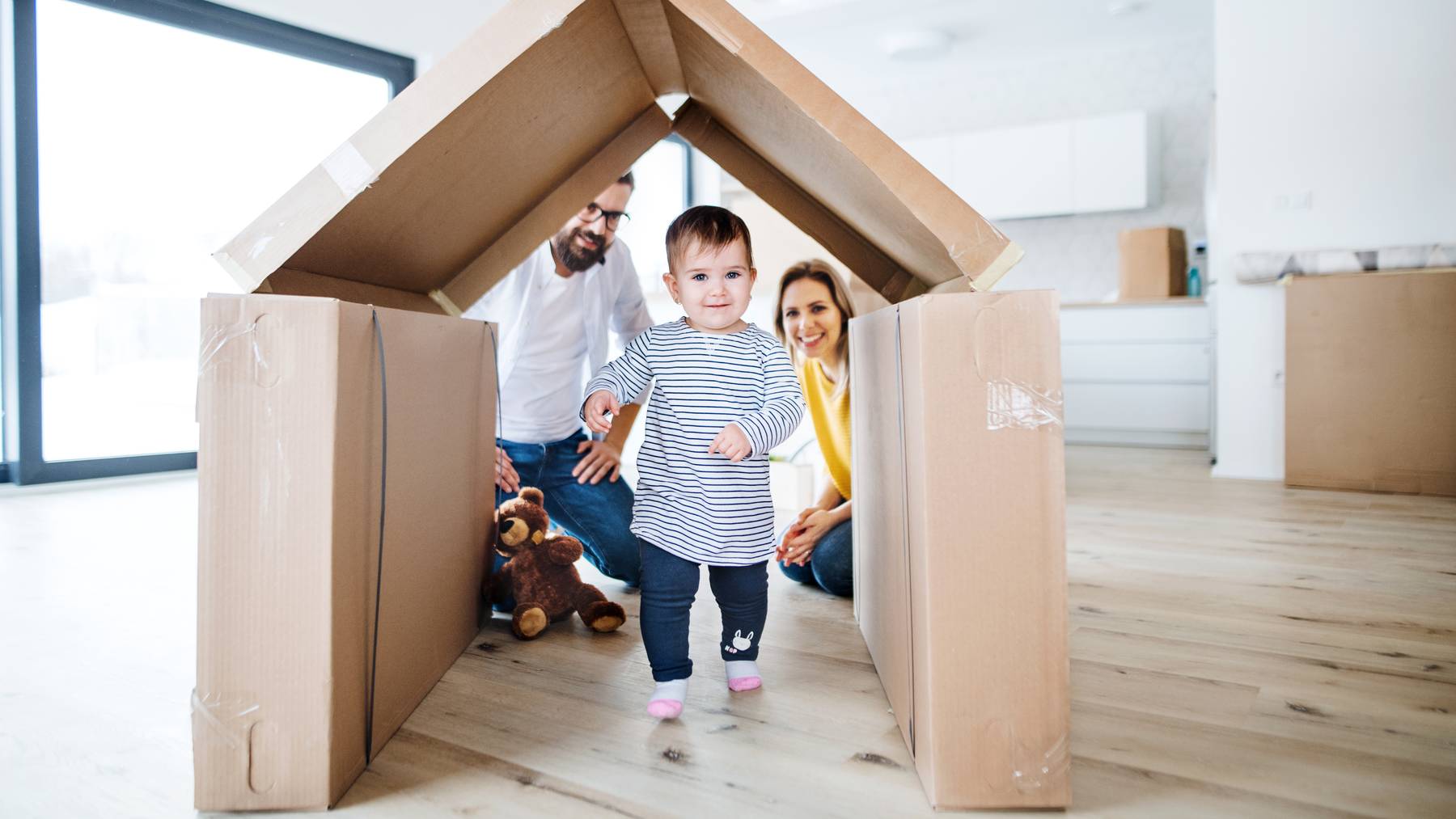Ein Kleinkind geht durch ein aus Kartons und Kisten gebautes Haus auf die Kamera zu. Dahinter sind die Eltern zu sehen.