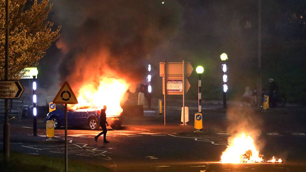 Ein Mann geht an einem brennenden Auto vorbei. Bei Protesten im nordirischen Belfast sind mehrere Polizisten verletzt worden. Foto: Peter Morrison/PA Wire/dpa
