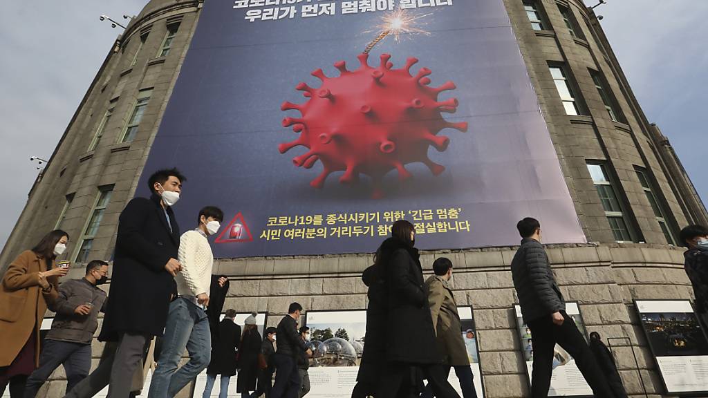 Menschen, die Gesichtsmasken tragen, gehen vor dem Rathaus von Seoul unter einem Transparent, das auf eine verstärkte soziale Distanzierungskampagne hinweist vorbei. Foto: Ahn Young-Joon/AP/dpa