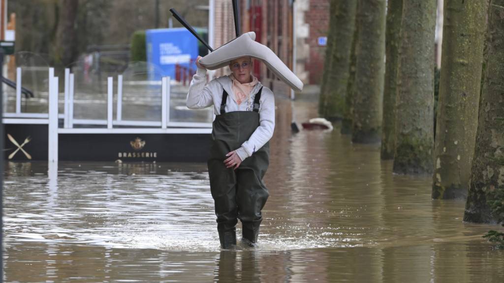 Eine Frau trägt einen Stuhl, während sie durch eine überflutete Straße watet. Foto: Matthieu Mirville/AP/dpa
