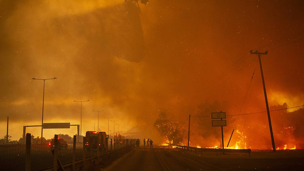 In der griechischen Kleinstadt Afidnes stehen Feuerwehrleute auf einer Straße und reinigen einen Feuerwehrschlauch, während im Hintergrund ein Hubschrauber Wasser über einem Waldbrand abwirft. Die dramatischen Waldbrände in Griechenland und der Türkei dauern unvermindert an. Foto: Angelos Tzortzinis/dpa