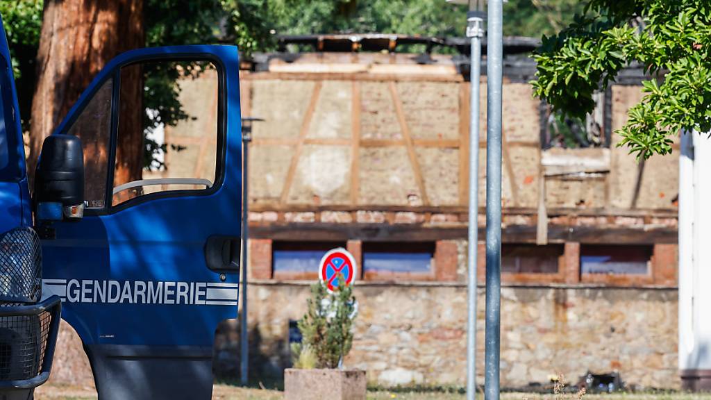Die Tür eines Fahrzeugs der Gendarmerie steht offen, während im Hintergrund ein Haus zu sehen ist, in dem am Vortag bei einem Feuer elf Menschen ums Leben kamen. Nach dem Brand in der Ferienunterkunft im Elsass dauern die Ermittlungen zur Brandursache an. Foto: Philipp von Ditfurth/dpa