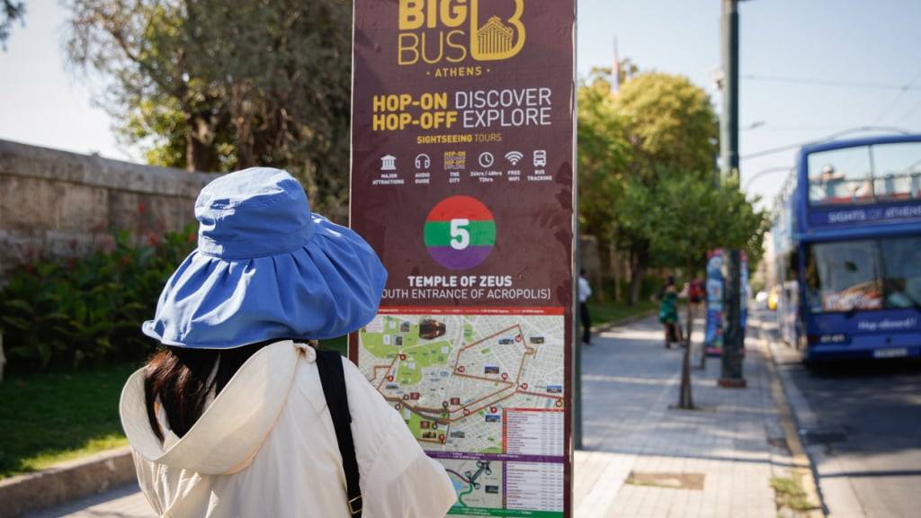 Eine Touristin mit Sonnenhut steht an einer Haltestelle für Sightseeing-Busse nahe der Akropolis. Foto: Socrates Baltagiannis/dpa