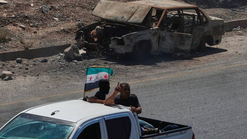 Kämpfer fahren auf der Ladefläche eines Pickups mit der syrischen Oppositionsflagge an einem ausgebrannten Auto vorbei auf der Autobahn von Daraa nach Damaskus. Foto: Omar Sanadiki/AP/dpa