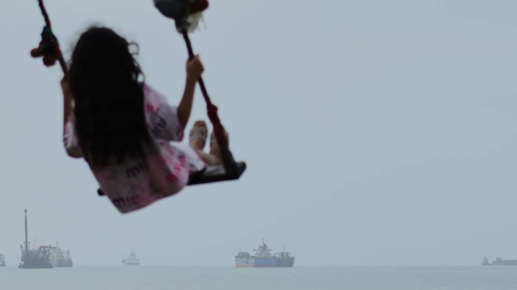 ARCHIV - Ein Mädchen schaukelt am Ufer, während Frachtschiffe und andere Handelsschiffe in der Straße von Hormus vor Anker liegen. Foto: Razieh Poudat/ISNA/AP/dpa