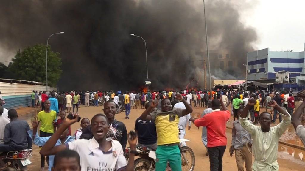Mit dem brennenden Hauptquartier der Regierungspartei im Rücken demonstrieren Anhänger meuternder Soldaten. Foto: Fatahoulaye Hassane Midou/AP/dpa